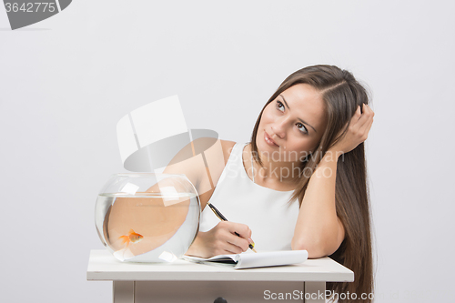 Image of Girl thoughtfully writing in a notebook, standing next to an aquarium with goldfish