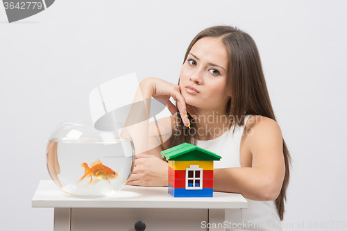 Image of Thoughtful girl sitting at a table on which there is an aquarium with goldfish and toy house