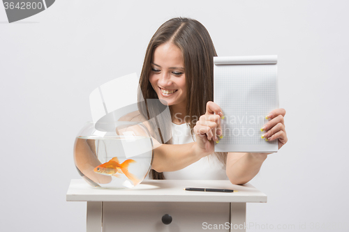 Image of Girl shows a notebook with a record, standing next to an aquarium with goldfish