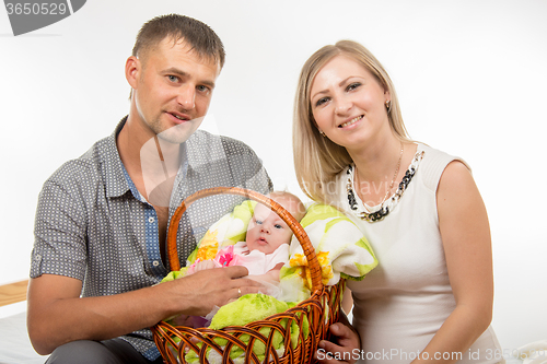 Image of Mom and dad sit on the bed and holding a two-month baby girl in a basket