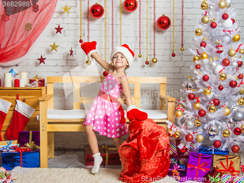 Image of Girl in a cap and mittens of Santa Claus dancing around the bag with Christmas gifts