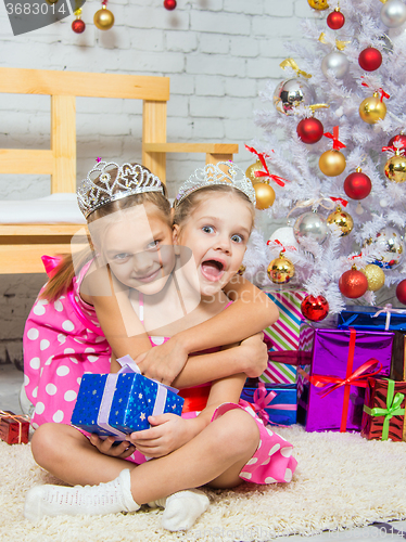 Image of Girl hugging another girl sitting on a rug with a gift at Christmas tree