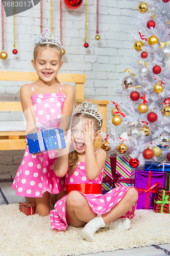 Image of The girl gives a gift to his sister sitting on the rug in front of the Christmas tree