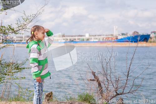 Image of A girl stands near the river on which the floating ship and looks into the distance