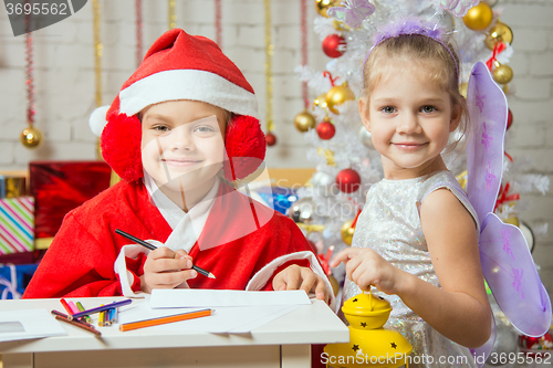Image of Girl dressed as Santa Claus writing a letter, standing next to a fairy with a flashlight in his hand