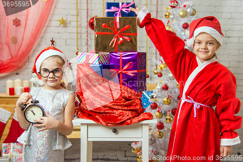 Image of A girl holding a clock, a girl dressed as Santa Claus is standing at the bag with gifts