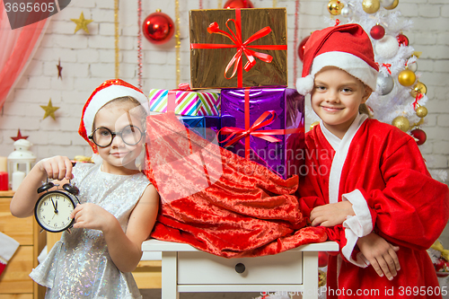 Image of Girl shows on the clock, a girl dressed as Santa Claus is standing at the bag with gifts