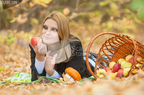 Image of Young beautiful girl lies on the on the foliage in autumn forest and looking at an apple in her hand