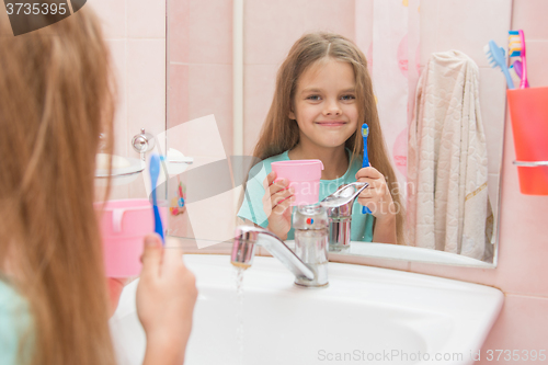 Image of Happy little girl standing with toothbrush and cup and looks in the mirror