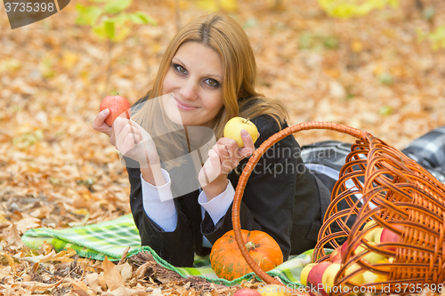 Image of Young girl lying on a rug in the autumn forest and holding two apples in the hands of