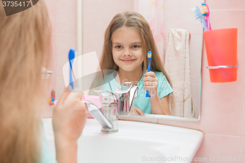 Image of  Six year old girl holding a toothbrush and looks at himself in the mirror, while in the bathroom