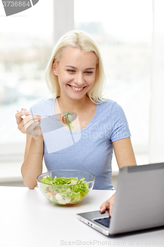 Image of smiling woman with laptop eating salad at home