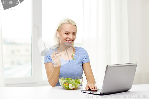 Image of smiling woman with laptop eating salad at home