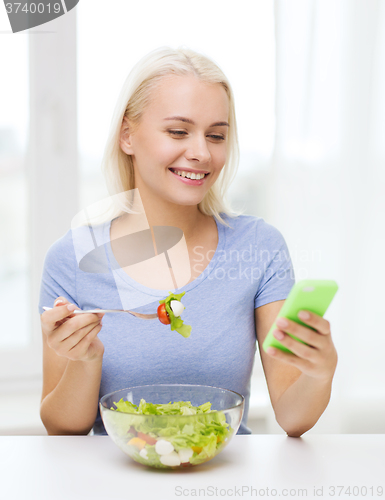 Image of smiling woman with smartphone eating salad at home