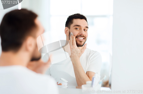 Image of happy young man applying cream to face at bathroom