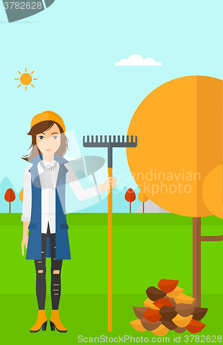 Image of Woman with rake standing near tree and heap of autumn leaves.