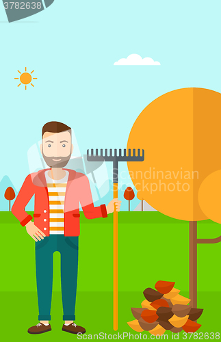 Image of Man with rake standing near tree and heap of autumn leaves.