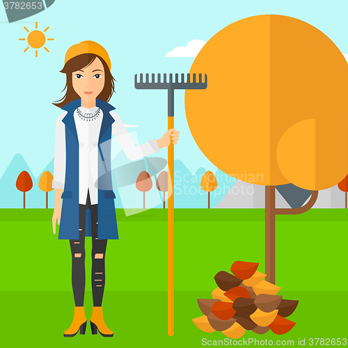 Image of Woman with rake standing near tree and heap of autumn leaves.