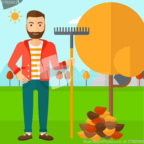 Image of Man with rake standing near tree and heap of autumn leaves.