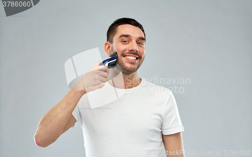 Image of smiling man shaving beard with trimmer over gray