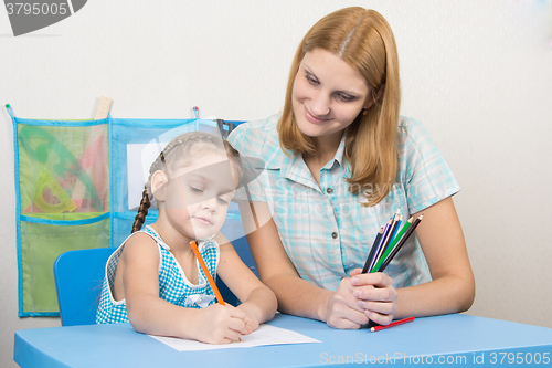 Image of The five-year child draws on a sheet of paper, next to the teacher holds pencils