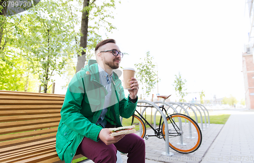 Image of happy hipster man eating sandwich with coffee