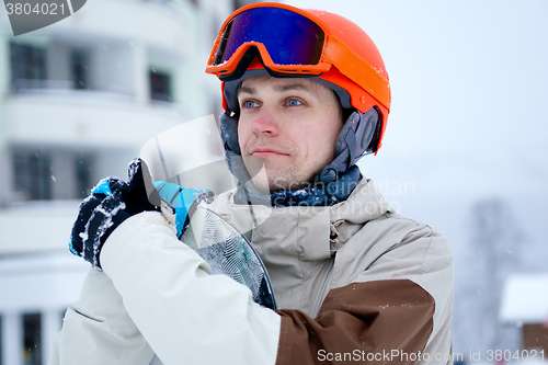 Image of Man snowboarder wearing orange helmet, grey jacket, black and blue gloves standing with snowboard