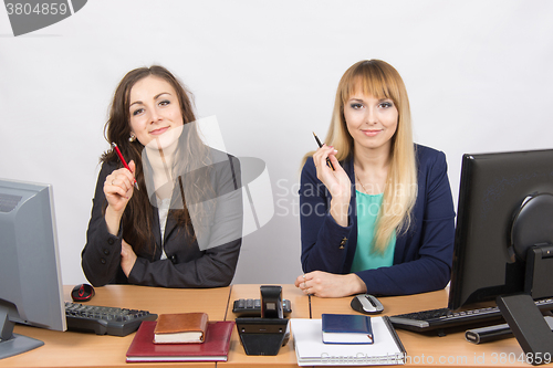 Image of Two business women sitting at a desk, holding pen in hand and look to the Frame