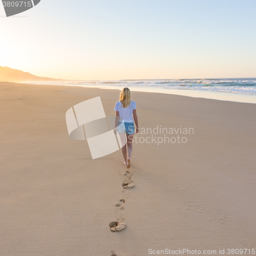 Image of Lady walking on sandy beach in sunset leaving footprints behind.