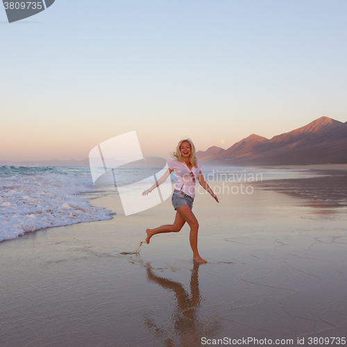 Image of Lady enjoying running from waves on sandy beach in sunset.