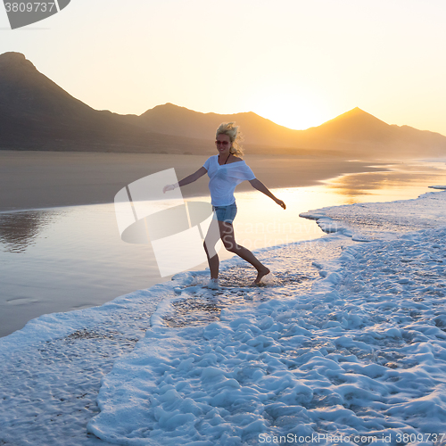 Image of Lady enjoying running from waves on sandy beach in sunset.