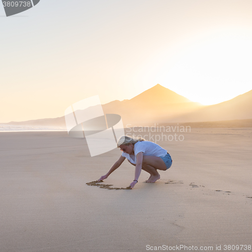 Image of Lady drawing heart shape in sand on beach.