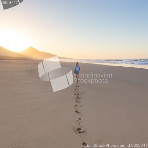 Image of Lady walking on sandy beach in sunset leaving footprints behind.