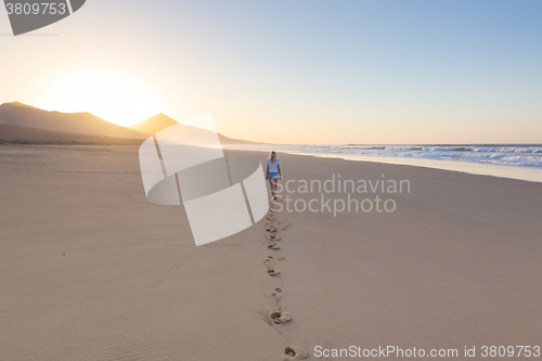 Image of Lady walking on sandy beach in sunset leaving footprints behind.