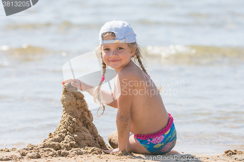 Image of Five-year girl in a cap sits on the sand on the beach and building sand castle