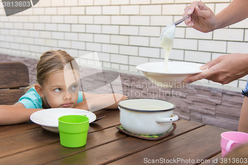 Image of Upset six-year girl dissatisfied looks at his mother who puts porridge for breakfast
