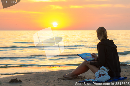 Image of Young girl sitting on the beach, drawing on a sheet of watercolor sunset and looked at the setting sun