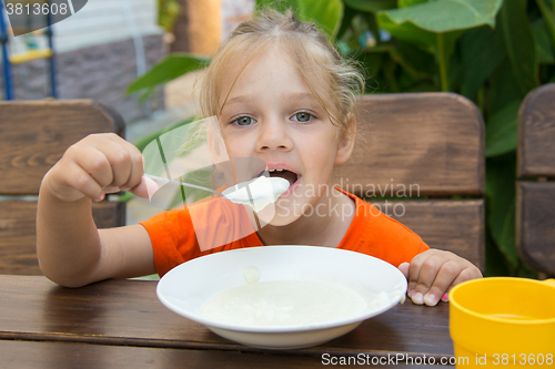 Image of Funny five-year girl with pleasure eats porridge for breakfast