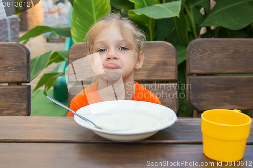 Image of Upset five year old girl poses faces unwilling to eat porridge for breakfast