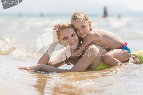 Image of Mom and sits on her back the baby lying in the water on the sandy beach and happily look into the frame