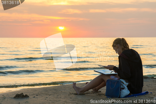 Image of Young girl sitting on the beach and draw on a sheet of watercolor sunset