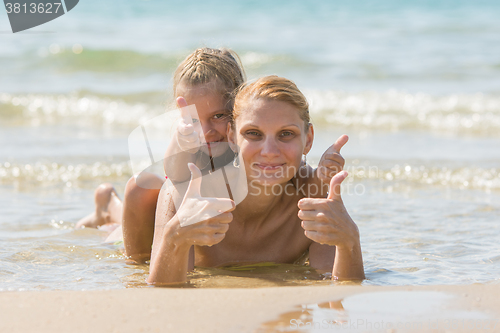Image of Mum with a daughter on his back lying on the beach and show. Girl and woman show thumbs up