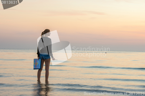 Image of Young girl standing in the water on the beach with a landscape painted on a sheet of paper and brush