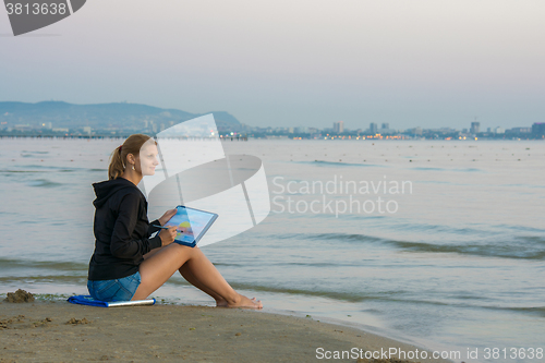 Image of Young girl sitting on the beach and drawing landscape with sunset