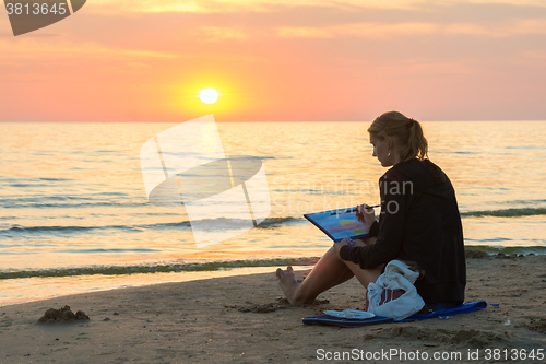 Image of  Young girl sitting on the beach and draw on a sheet of watercolor sunset