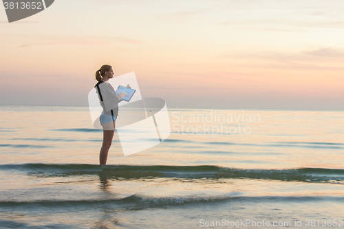 Image of Young girl standing in the water on the beach, and draws on the colors of the sunset on a piece of paper