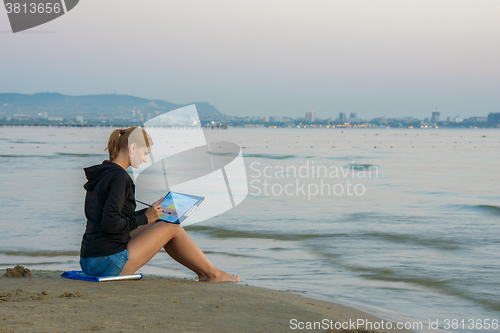 Image of The young artist is sitting on the beach and drawing landscape with sunset