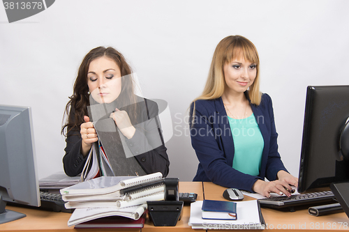 Image of Female colleagues in the office, one asleep on a folder, the other happy working on a computer