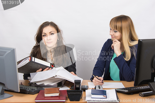 Image of The situation in the office - one employee inundated with work and looking at the frame, the other does nothing, and with a smile looking at her
