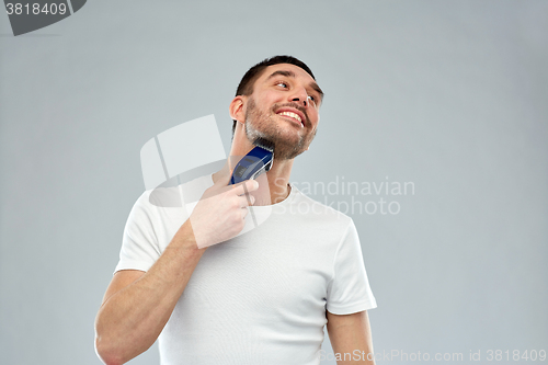 Image of smiling man shaving beard with trimmer over gray